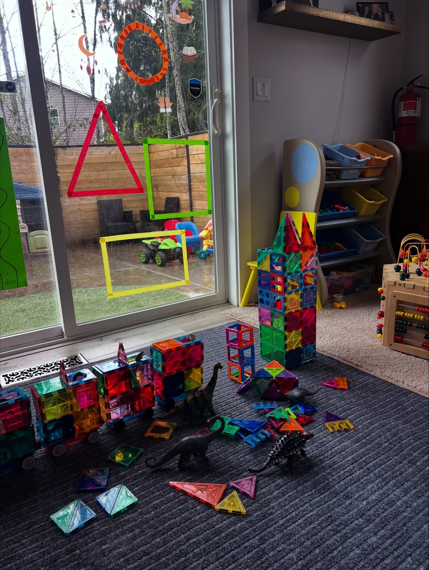 A corner of the playroom with magnetic tiles, dinosaur figures, and a window onto the yard